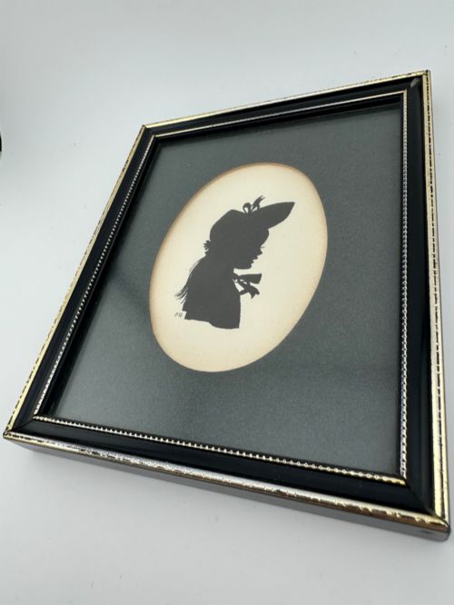 fig 6. A framed Art Deco silhouette portrait depicting a lady in profile with a decorative hat inside an oval cream mount surrounded by a black and gold frame.