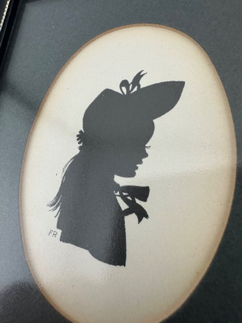 fig 7. A framed Art Deco silhouette portrait depicting a lady in profile with a decorative hat inside an oval cream mount surrounded by a black and gold frame.