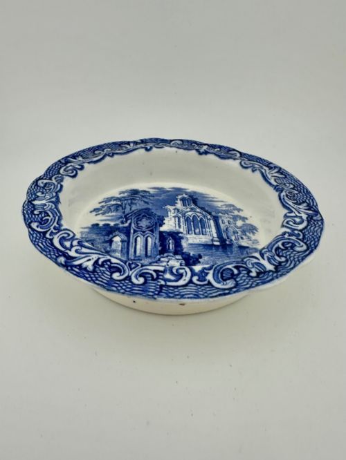 fig 1. A circular blue and white transferware ceramic dish featuring a central printed depiction of abbey ruins framed by a decorative scrolled border.