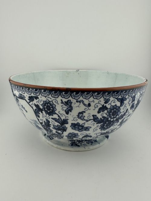 fig 1. A deep circular footed earthenware bowl decorated with blue and white floral and landscape transferware patterns with a brown rim.
