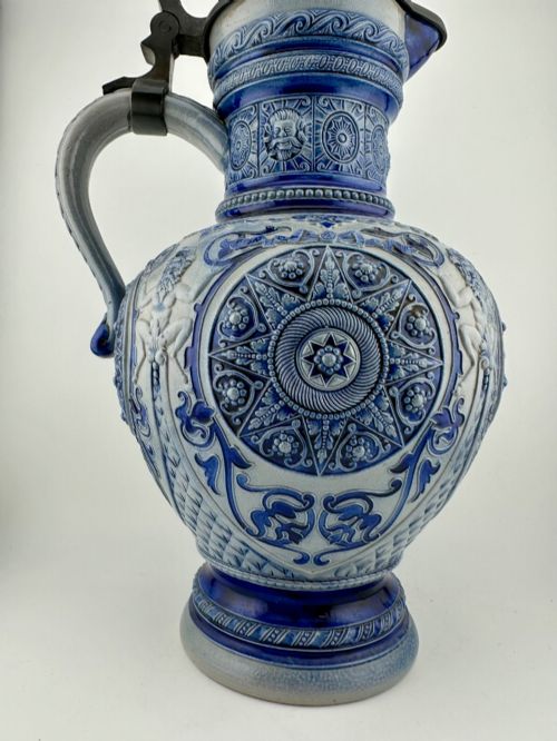 fig 1. A blue and grey salt-glazed stoneware jug featuring relief-moulded geometric patterns, mask motifs, and a hinged pewter lid.