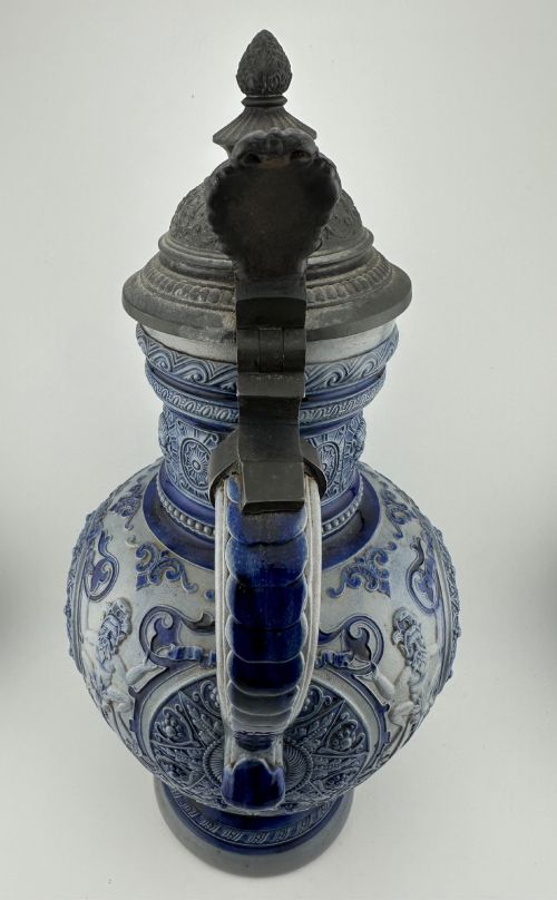 fig 3. A blue and grey salt-glazed stoneware jug featuring relief-moulded geometric patterns, mask motifs, and a hinged pewter lid.