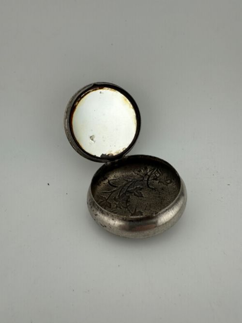 fig 1. A circular antique Art Nouveau white metal compact box featuring a relief-carved iris floral design with brass highlights and a textured background.