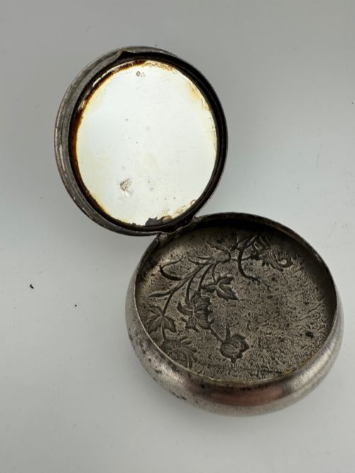 fig 2. A circular antique Art Nouveau white metal compact box featuring a relief-carved iris floral design with brass highlights and a textured background.