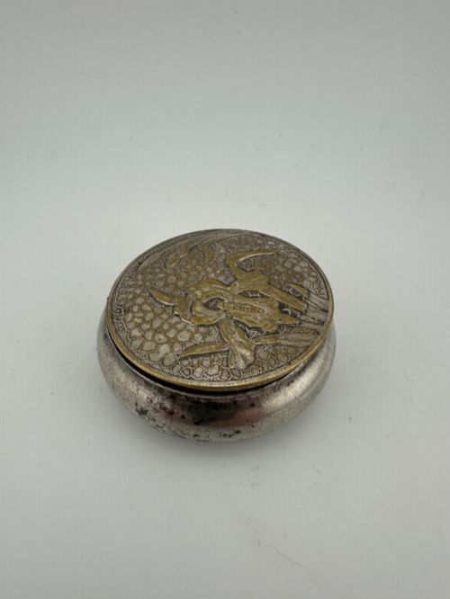 fig 3. A circular antique Art Nouveau white metal compact box featuring a relief-carved iris floral design with brass highlights and a textured background.