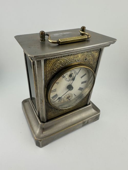fig 1. An Edwardian carriage clock with a silvered metal casing, brass carrying handle, and engraved brass mask around a white enamel dial.