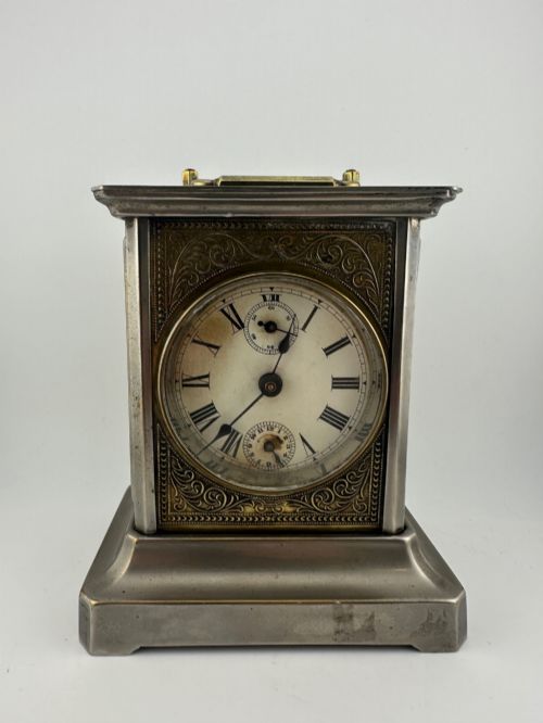fig 2. An Edwardian carriage clock with a silvered metal casing, brass carrying handle, and engraved brass mask around a white enamel dial.
