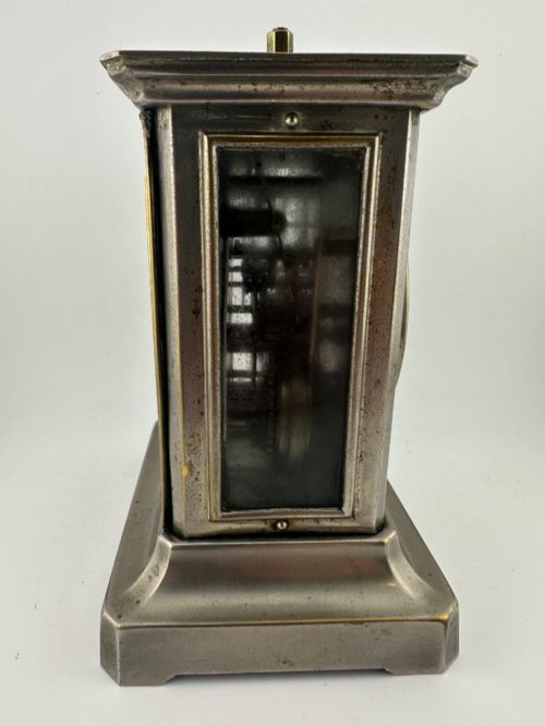 fig 3. An Edwardian carriage clock with a silvered metal casing, brass carrying handle, and engraved brass mask around a white enamel dial.