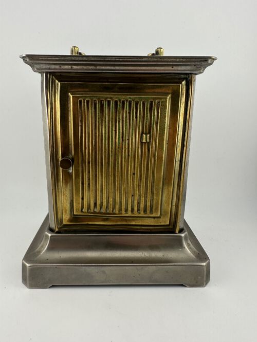 fig 4. An Edwardian carriage clock with a silvered metal casing, brass carrying handle, and engraved brass mask around a white enamel dial.