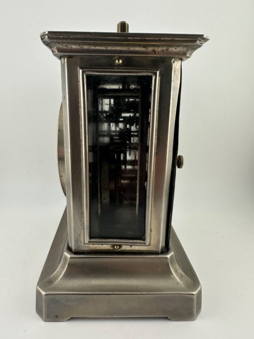 fig 5. An Edwardian carriage clock with a silvered metal casing, brass carrying handle, and engraved brass mask around a white enamel dial.