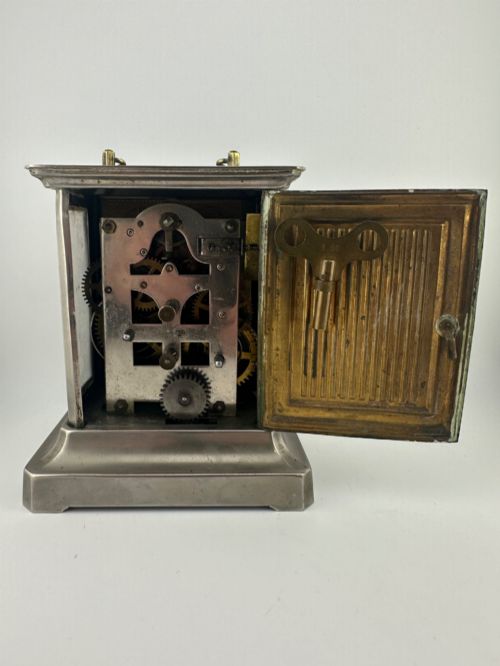 fig 6. An Edwardian carriage clock with a silvered metal casing, brass carrying handle, and engraved brass mask around a white enamel dial.