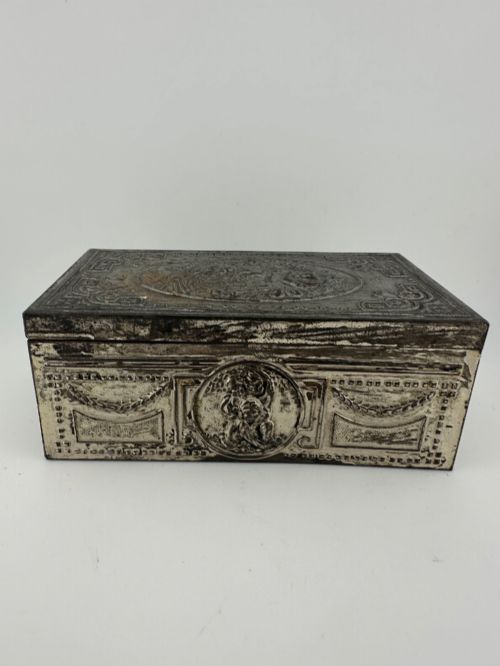 fig 1. A rectangular silver-grey metal box featuring embossed neoclassical cherub medallions and relief swags across its hinged lid and sides.