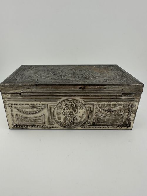 fig 3. A rectangular silver-grey metal box featuring embossed neoclassical cherub medallions and relief swags across its hinged lid and sides.