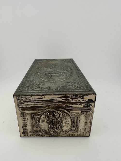 fig 4. A rectangular silver-grey metal box featuring embossed neoclassical cherub medallions and relief swags across its hinged lid and sides.