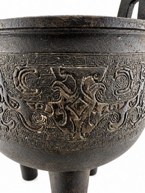fig 3. A dark patinated bronze tripod censer featuring upright loop handles and relief taotie mask carvings around the vessel body.