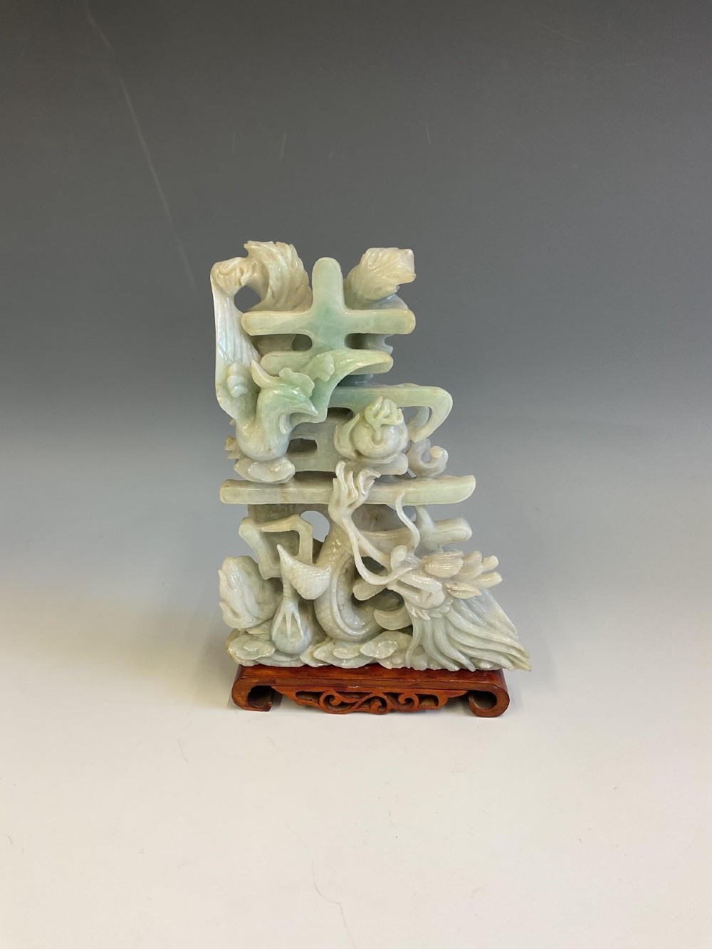 A pale green carved jade sculpture depicting a dragon, phoenix, and double happiness character mounted on a dark carved hardwood stand.