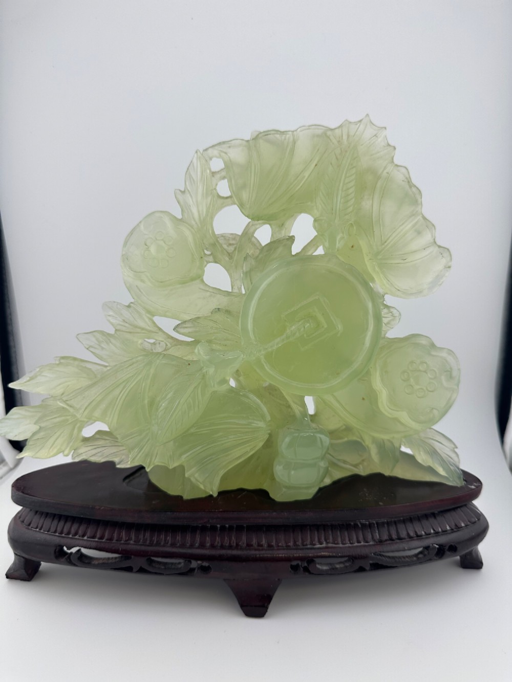 A carved pale green Xiuyan jade sculpture depicting a lotus flower, foliage, and a central cash coin, displayed on a dark carved wooden stand. View III