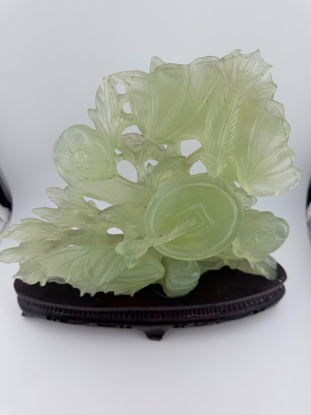 A carved pale green Xiuyan jade sculpture depicting a lotus flower, foliage, and a central cash coin, displayed on a dark carved wooden stand. View IV