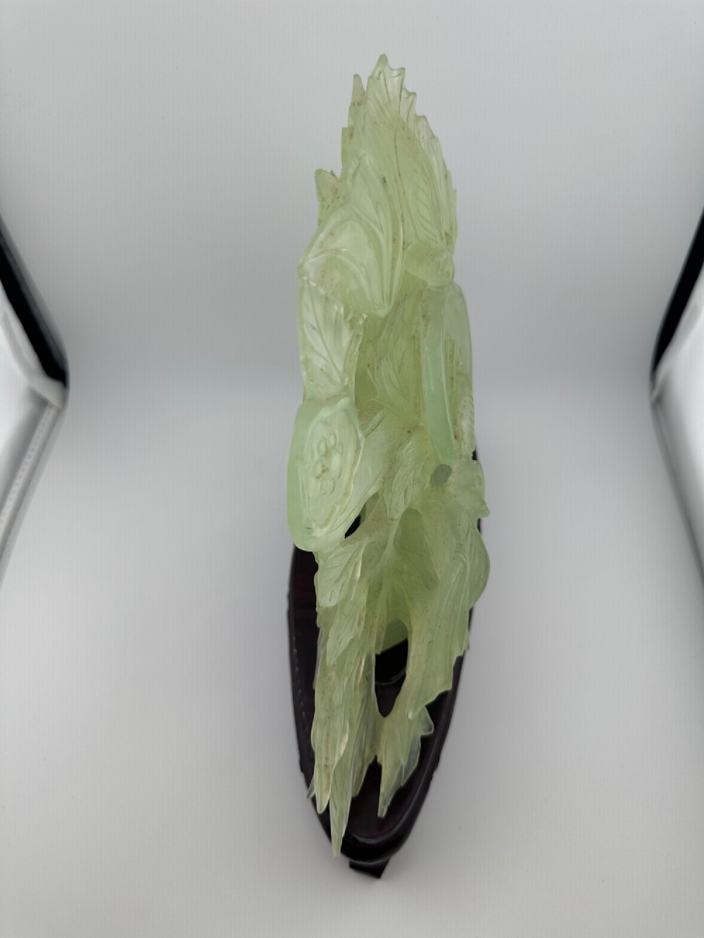 A carved pale green Xiuyan jade sculpture depicting a lotus flower, foliage, and a central cash coin, displayed on a dark carved wooden stand. View V