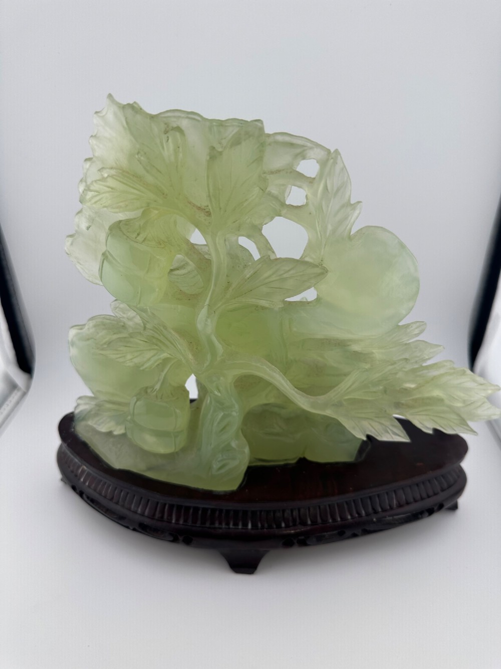 A carved pale green Xiuyan jade sculpture depicting a lotus flower, foliage, and a central cash coin, displayed on a dark carved wooden stand. View VI
