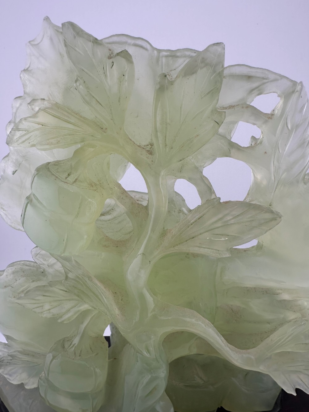 A carved pale green Xiuyan jade sculpture depicting a lotus flower, foliage, and a central cash coin, displayed on a dark carved wooden stand. View VII