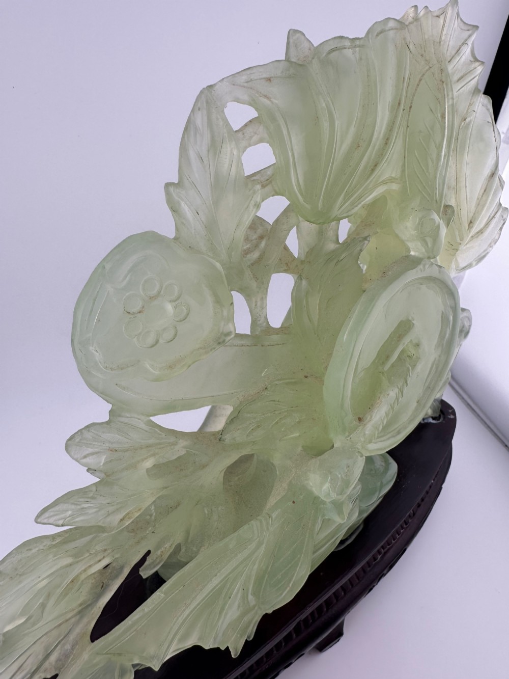 A carved pale green Xiuyan jade sculpture depicting a lotus flower, foliage, and a central cash coin, displayed on a dark carved wooden stand. View VIII