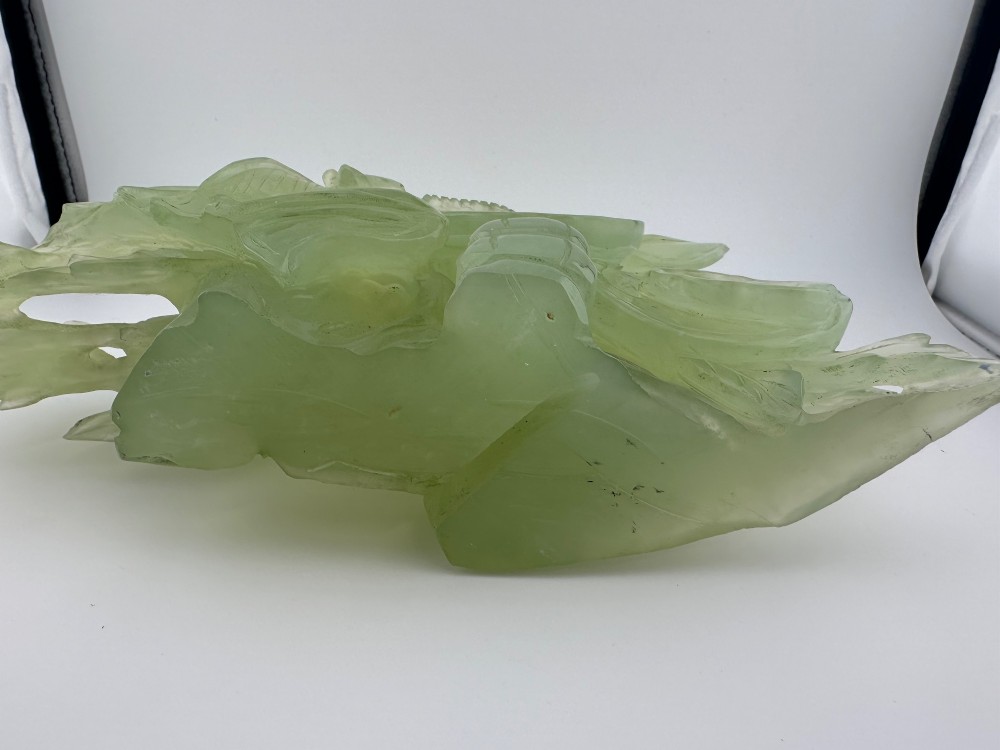 A carved pale green Xiuyan jade sculpture depicting a lotus flower, foliage, and a central cash coin, displayed on a dark carved wooden stand. View IX