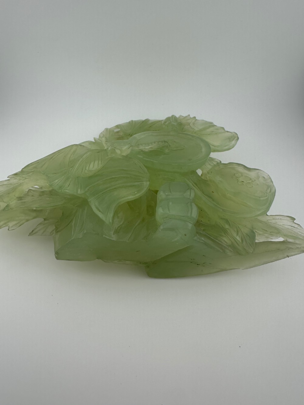 A carved pale green Xiuyan jade sculpture depicting a lotus flower, foliage, and a central cash coin, displayed on a dark carved wooden stand. View X