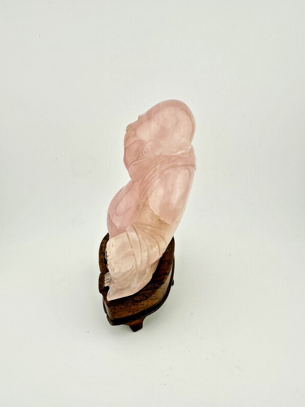 A hand-carved pink rose quartz figure of a seated Laughing Buddha resting upon a custom dark wood stand. View III