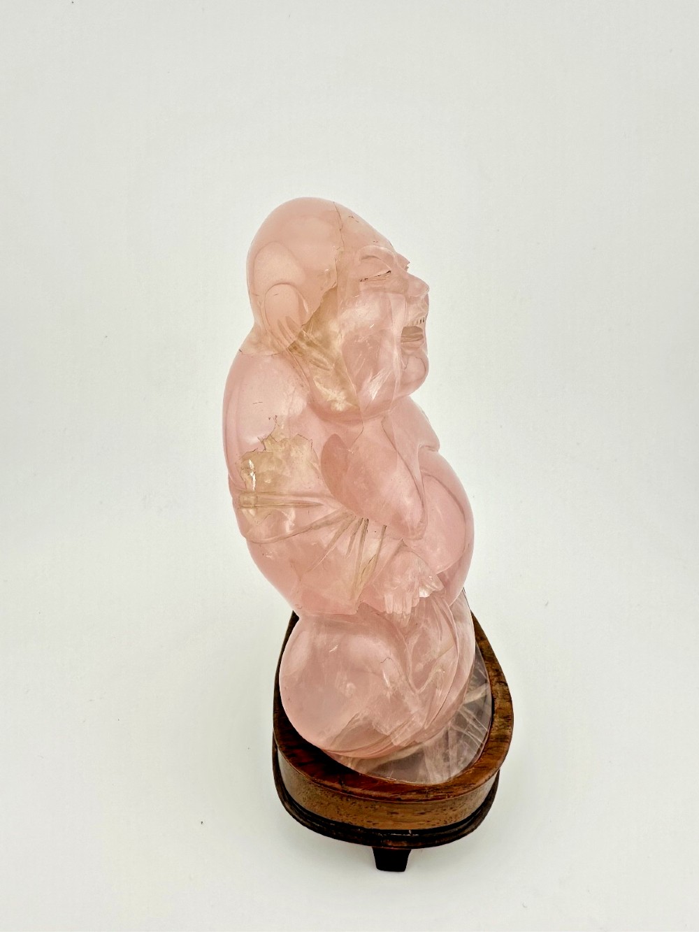 A hand-carved pink rose quartz figure of a seated Laughing Buddha resting upon a custom dark wood stand. View IV