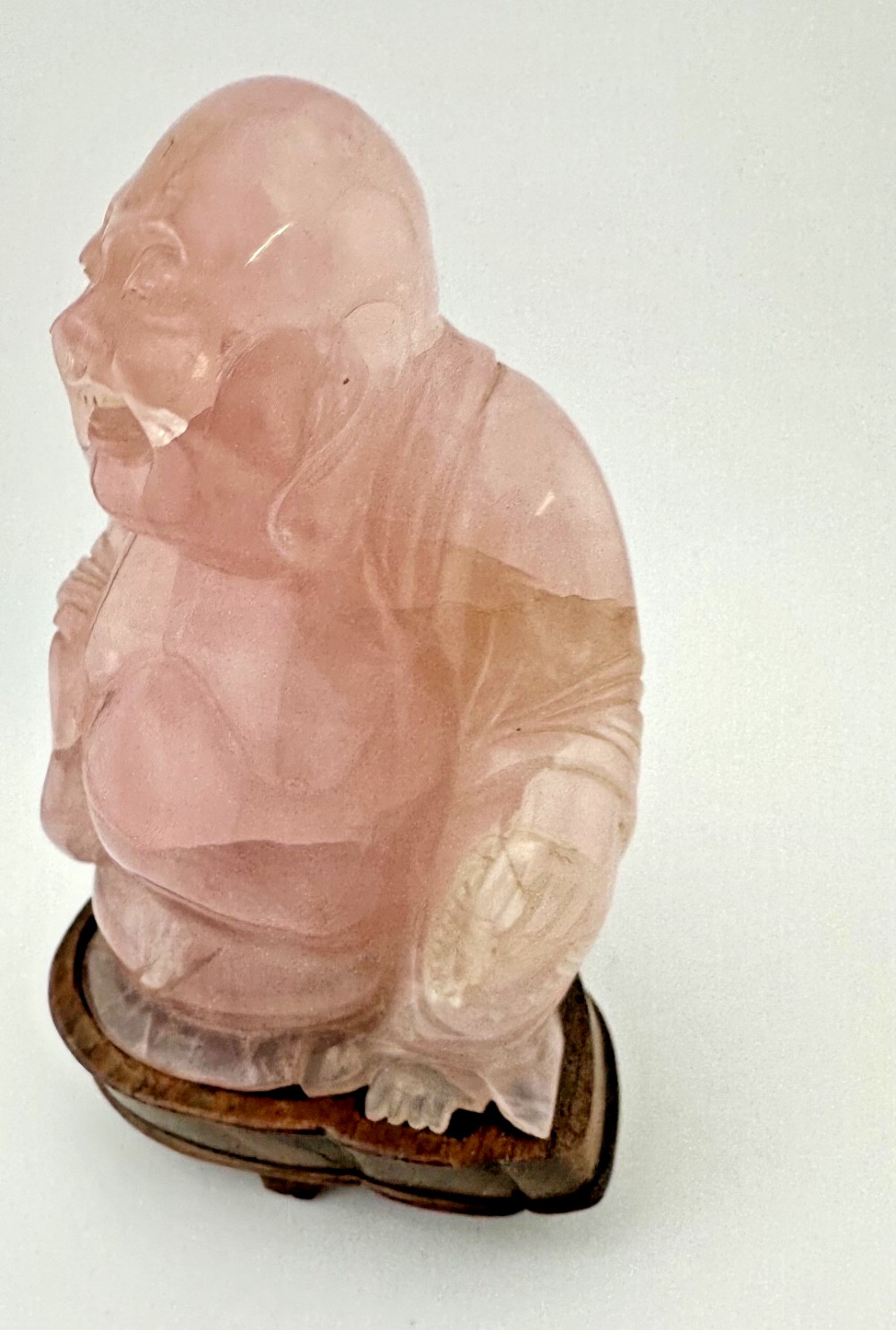 A hand-carved pink rose quartz figure of a seated Laughing Buddha resting upon a custom dark wood stand. View VII