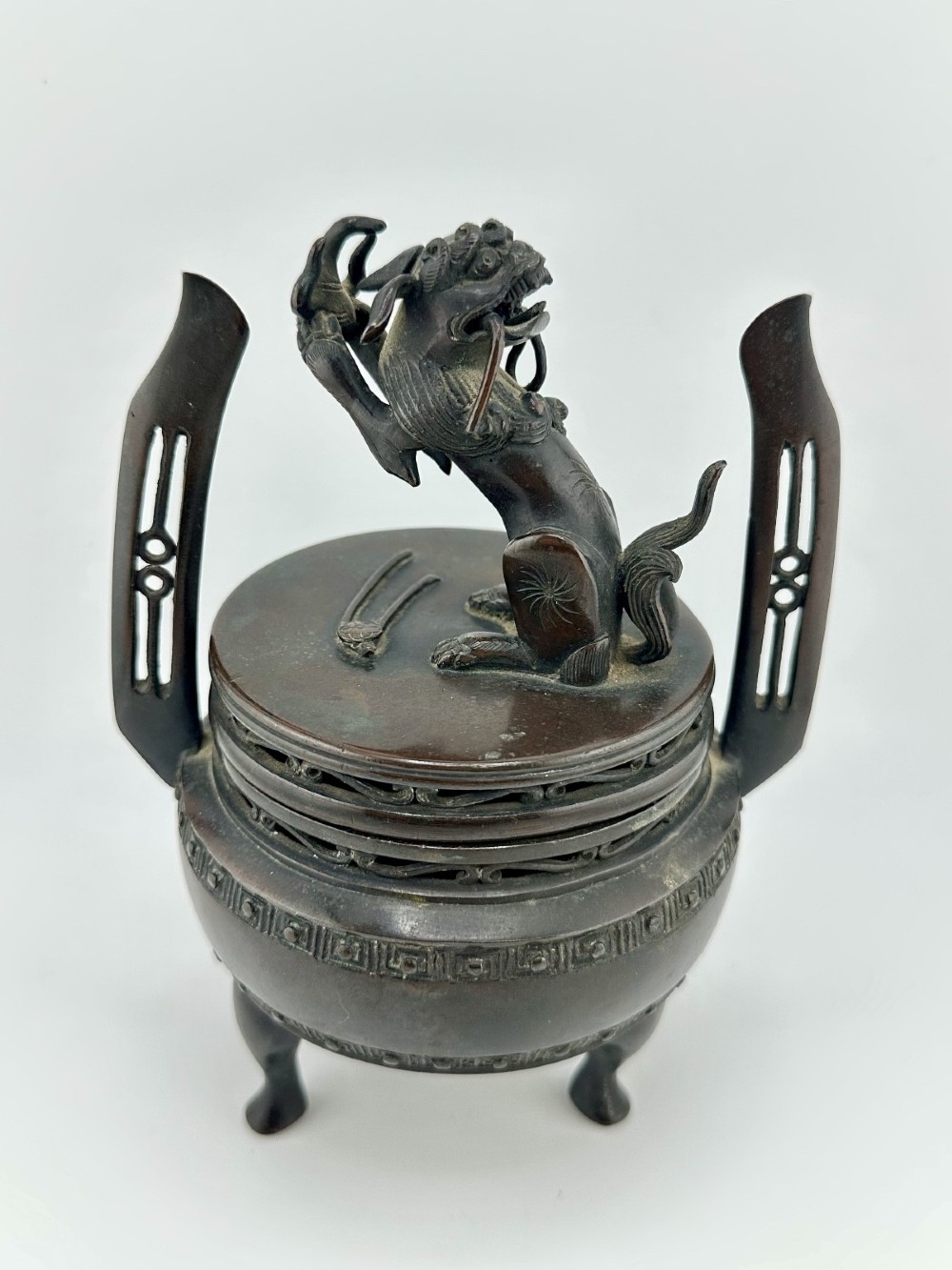 A dark patinated bronze tripod censer featuring raised loop handles, a decorative meander border, and a fitted lid with a standing Qilin finial.
