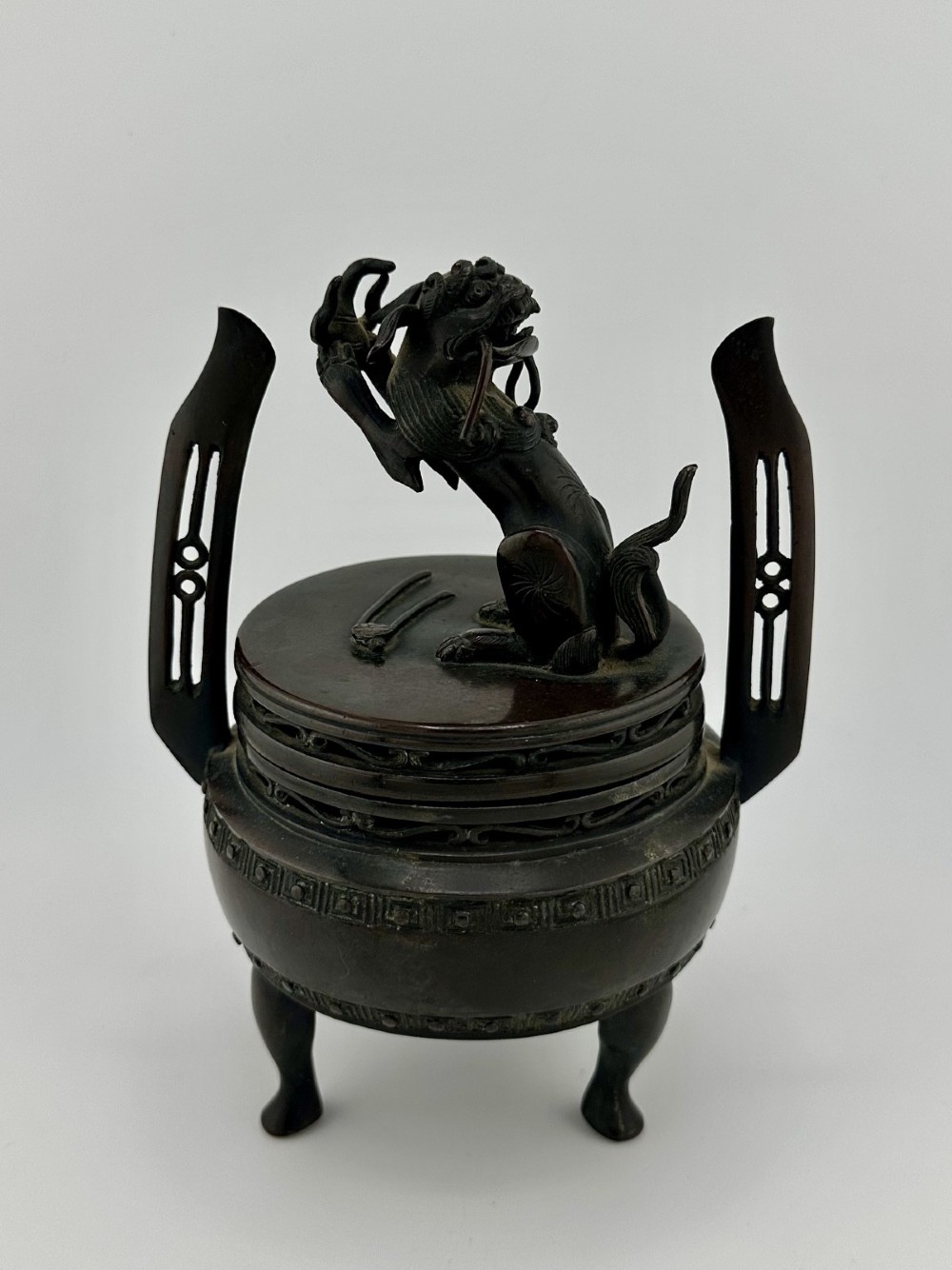 A dark patinated bronze tripod censer featuring raised loop handles, a decorative meander border, and a fitted lid with a standing Qilin finial. View II