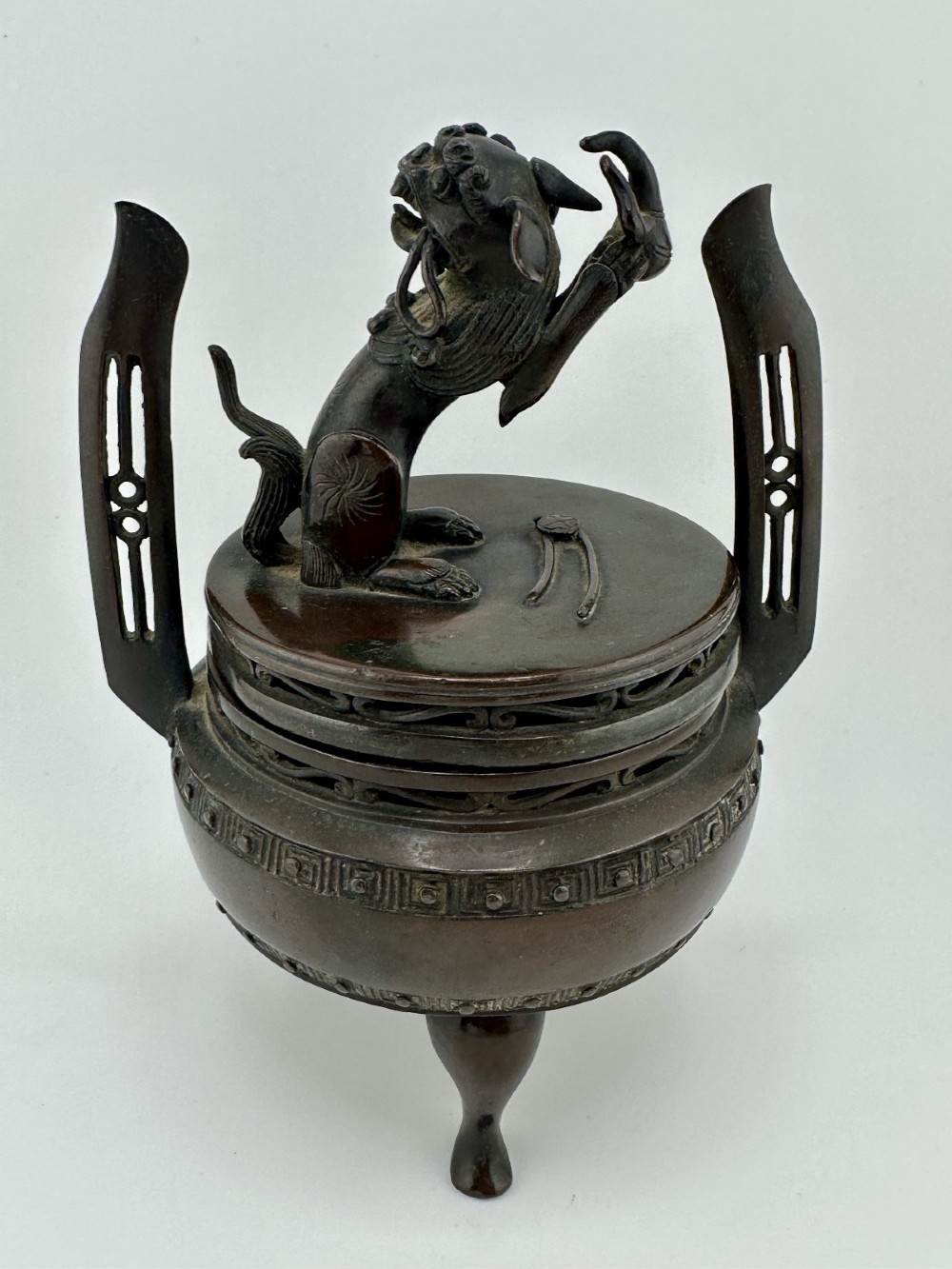 A dark patinated bronze tripod censer featuring raised loop handles, a decorative meander border, and a fitted lid with a standing Qilin finial. View VI