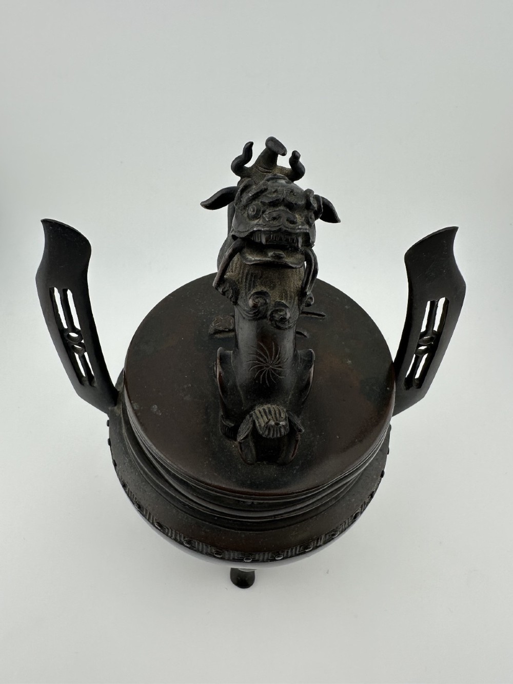 A dark patinated bronze tripod censer featuring raised loop handles, a decorative meander border, and a fitted lid with a standing Qilin finial. View XI
