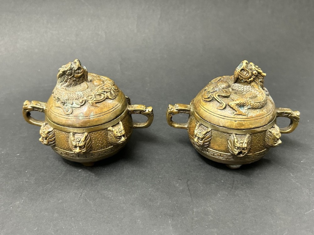 A pair of patinated bronze tripod censers featuring sculpted dragon finials on the lids, side handles, and relief mask carvings around the circular bodies.