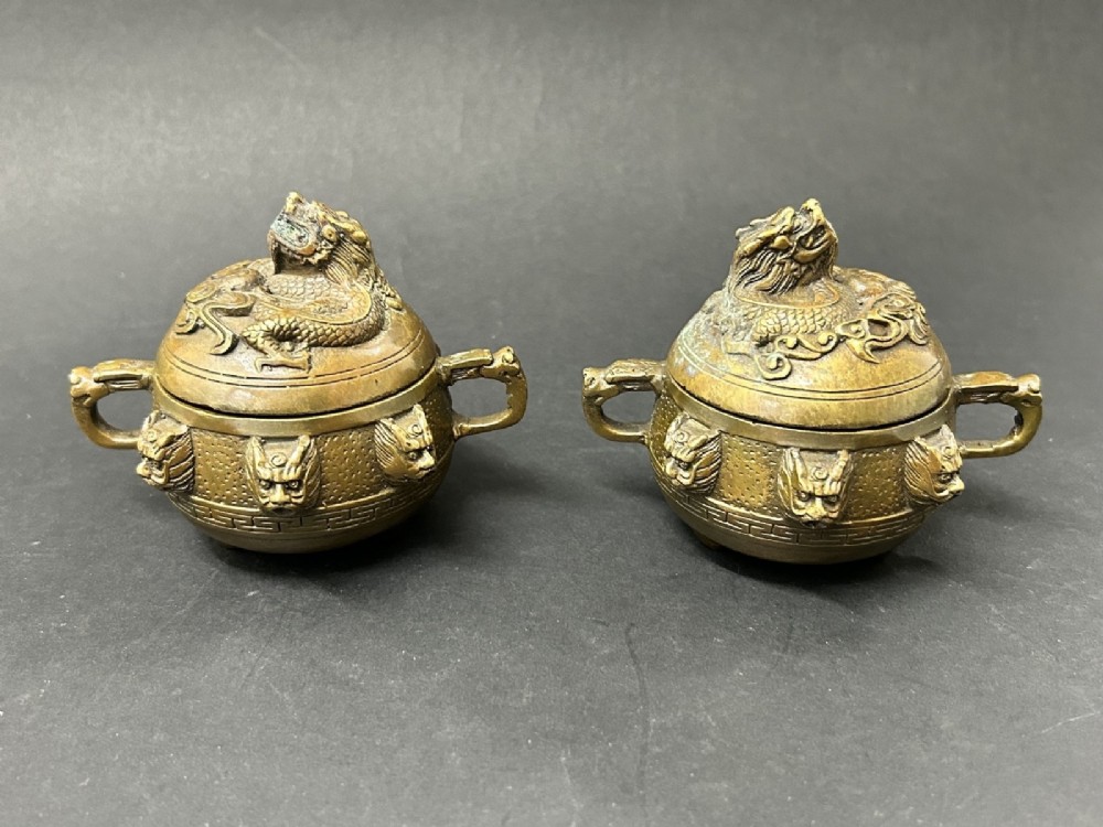 A pair of patinated bronze tripod censers featuring sculpted dragon finials on the lids, side handles, and relief mask carvings around the circular bodies. View II