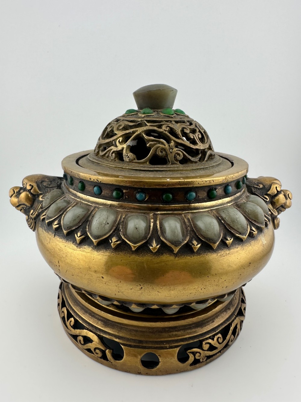 An antique gilded bronze Chinese censer featuring hardstone and jade cabochon inlays, beast-head handles, a pierced cover, and a matching carved stand.