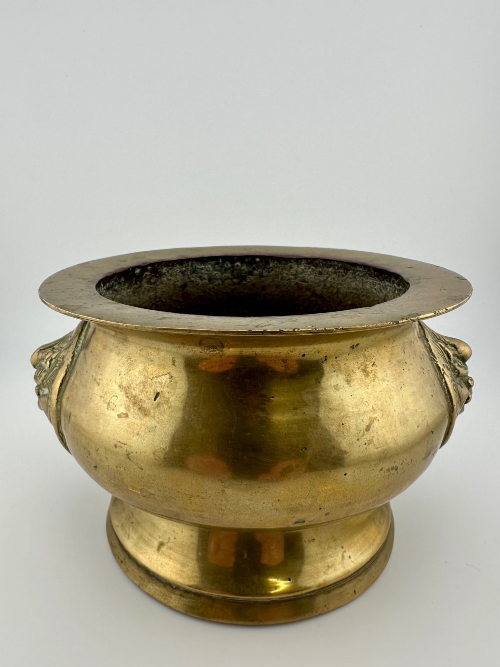 A rounded antique brass Chinese censer featuring a flared rim, pedestal base, and sculpted lion mask handles on either side.