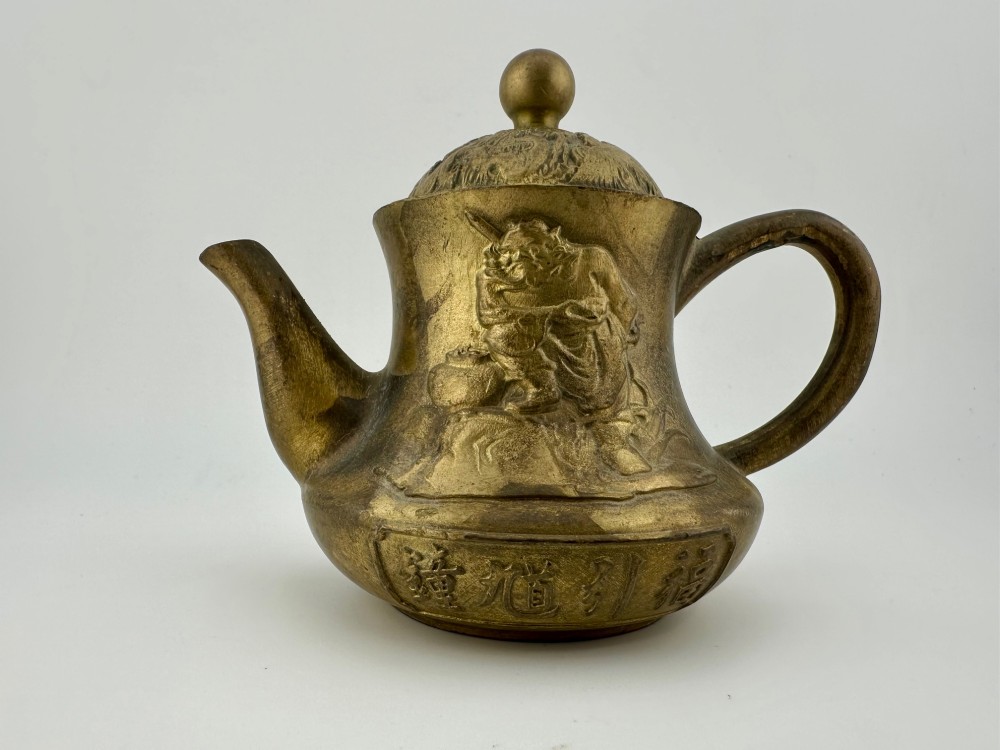 A cast brass Chinese teapot featuring raised relief figures, traditional Chinese characters along the base, a curved spout, handle, and a round lid knob.