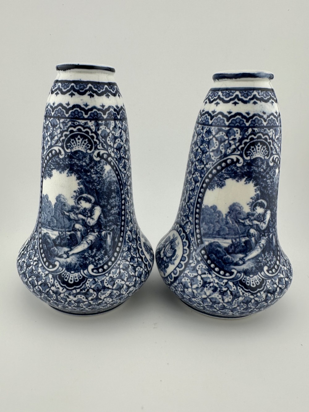 A pair of antique blue and white ceramic vases featuring central pastoral vignettes framed by elaborate scrollwork patterns.