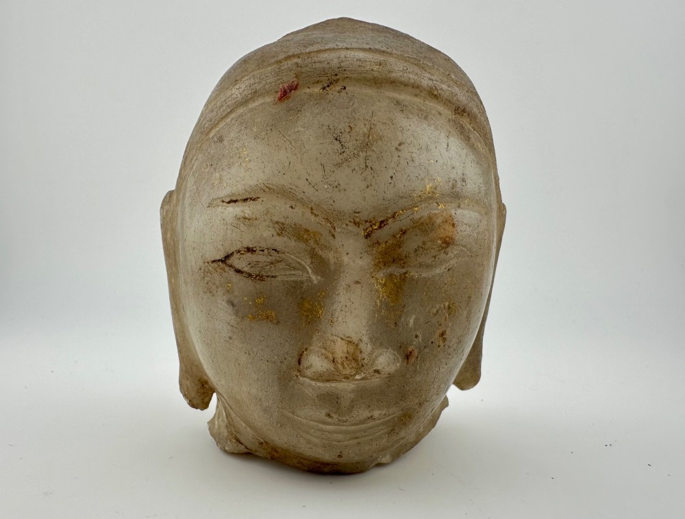 A carved off-white hardstone head fragment featuring facial features, elongated earlobes, and faint traces of gilding across the surface.