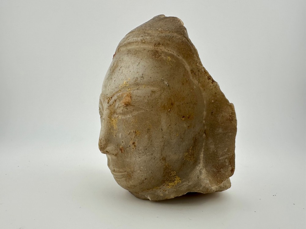 A carved off-white hardstone head fragment featuring facial features, elongated earlobes, and faint traces of gilding across the surface. View II