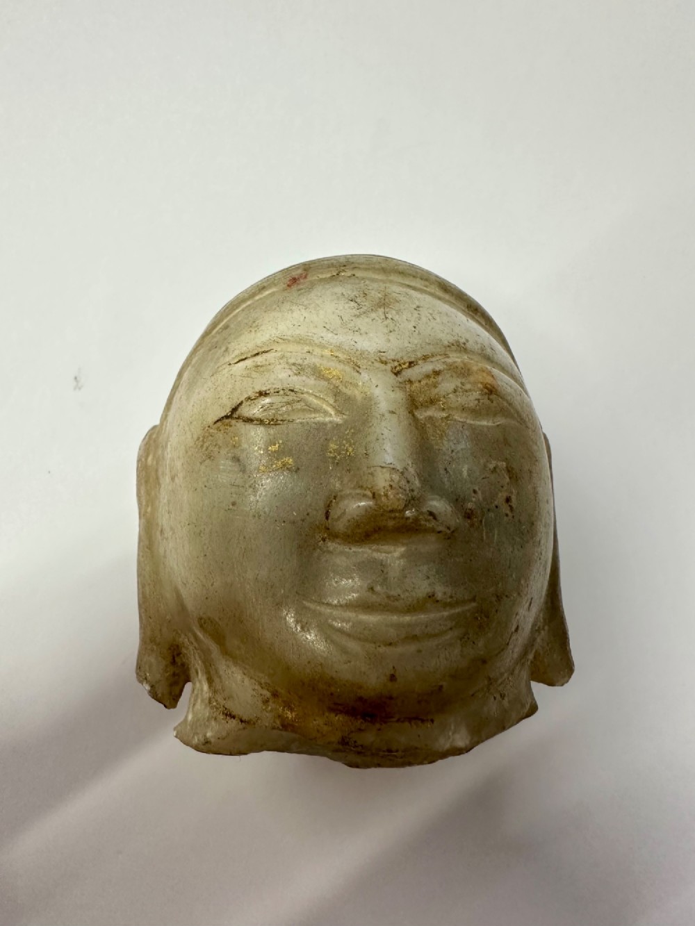 A carved off-white hardstone head fragment featuring facial features, elongated earlobes, and faint traces of gilding across the surface. View X