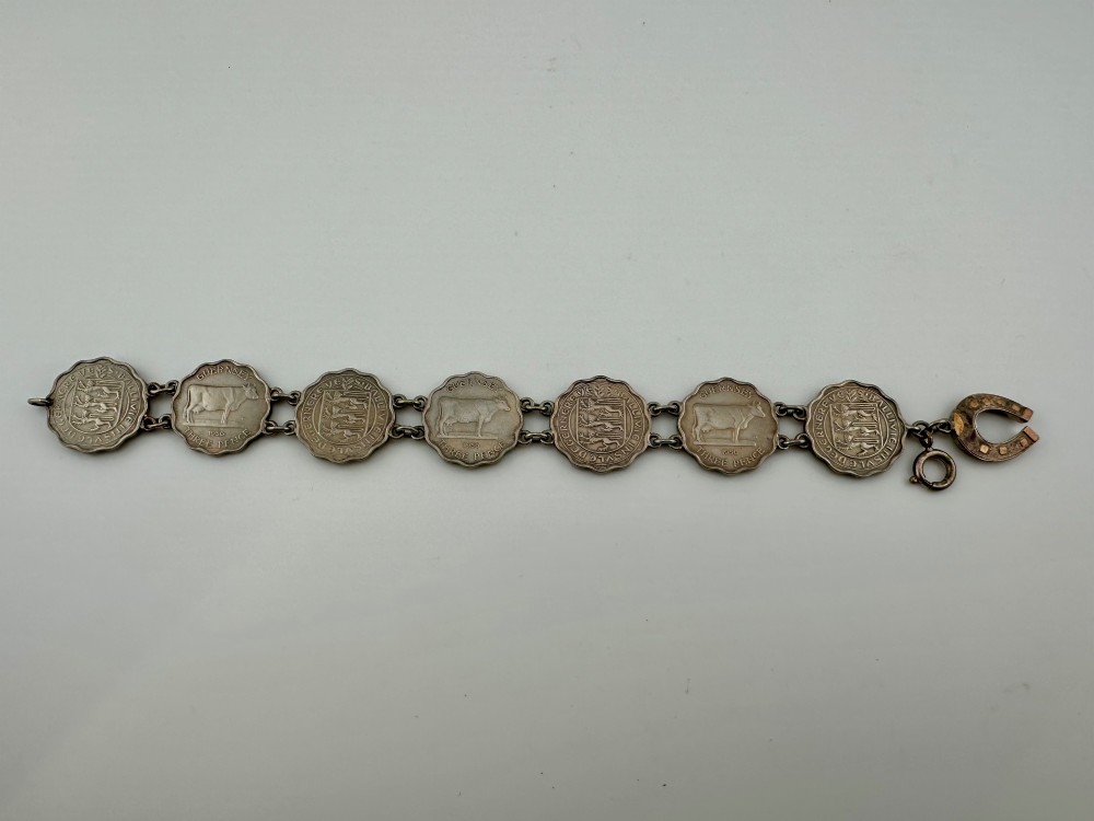 A silver-toned metal bracelet made from scalloped 1956 Guernsey threepence coins featuring a cow and coat of arms, connected to a horseshoe charm and bolt ring clasp. View II