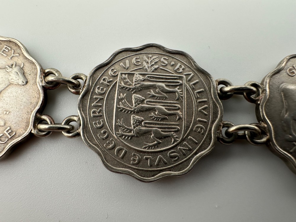A silver-toned metal bracelet made from scalloped 1956 Guernsey threepence coins featuring a cow and coat of arms, connected to a horseshoe charm and bolt ring clasp. View VI