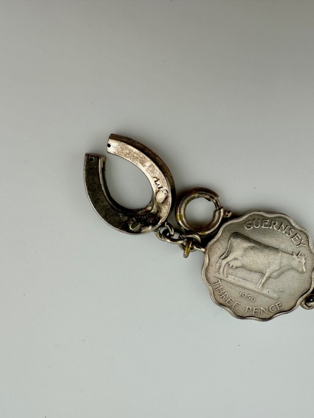 A silver-toned metal bracelet made from scalloped 1956 Guernsey threepence coins featuring a cow and coat of arms, connected to a horseshoe charm and bolt ring clasp. View VIII