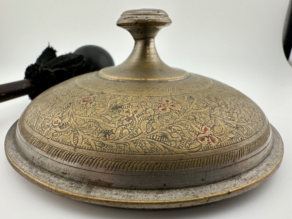 An antique domed Indian brass hand gong with detailed floral engravings, a turned central finial handle, and a dark wooden striker.