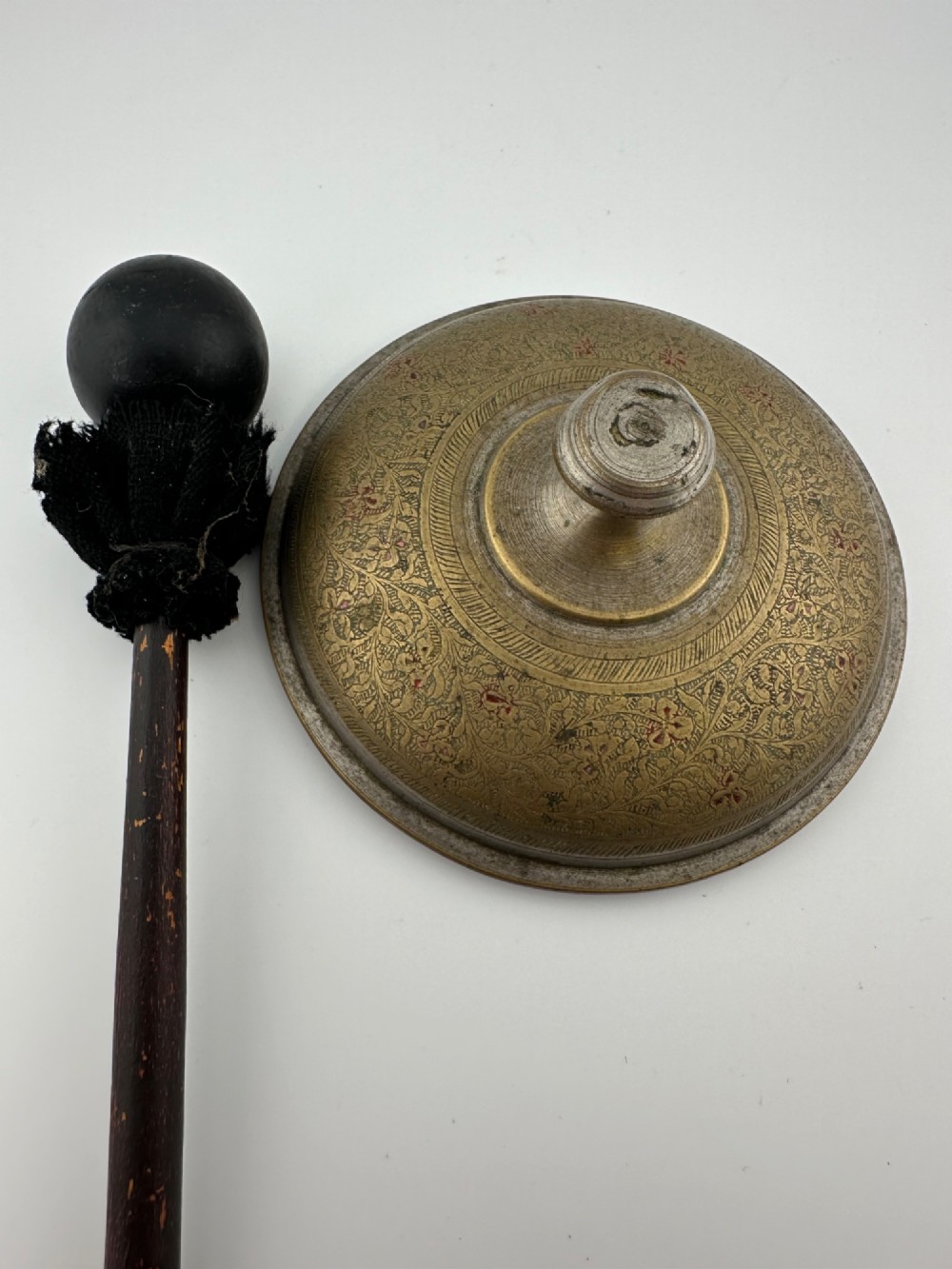An antique domed Indian brass hand gong with detailed floral engravings, a turned central finial handle, and a dark wooden striker. View II