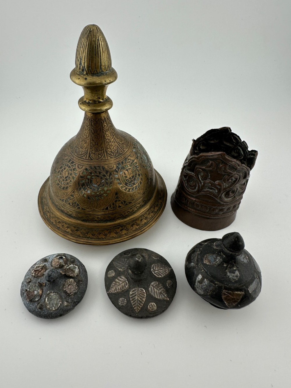 A five-piece group of antique Persian Qajar metalware, including an engraved brass incense burner lid with a finial, a dark carved vessel collar, and three small circular lids.